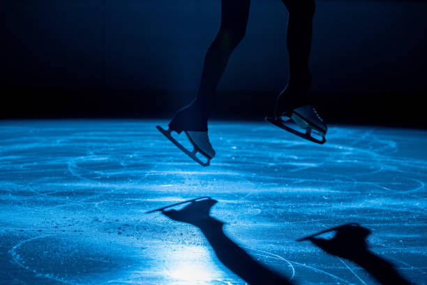 ジャンプの瞬間のフィギュアスケートの女性アスリート。フィギュアスケート用の白いスケートを履いた女性の足。青い光のある暗い氷のアリーナでスケートスキルを練習する。クローズア� - フィギュアスケート ストックフォトと画像