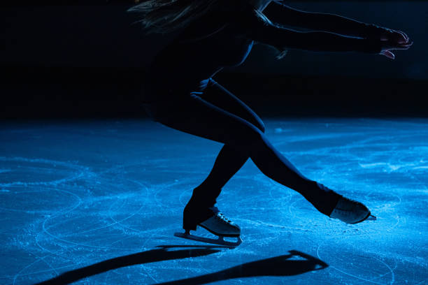 la mujer de patinaje artístico lleva ropa deportiva negra y patina en la pista de hielo, entrenando por la noche en los rayos de luz azul. mujer joven preparándose para la competencia, entrenando la rotación y las habilidades de deslizamiento. cerrar - patinaje artístico fotografías e imágenes de stock