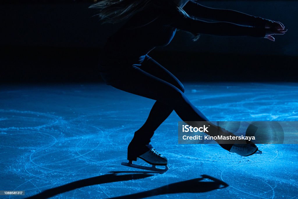 Figure skating lady is wearing black sportswear is skating on ice rink, training at night in the rays of blue light. Young woman preparing to competition, training rotation and slides skills. Close up Figure skating lady is wearing black sportswear is skating on ice rink, training at night in the rays of blue light. Silhouette of young woman preparing to competition, training rotation and slides skills. Details of legs in skates close up. Figure Skating Stock Photo