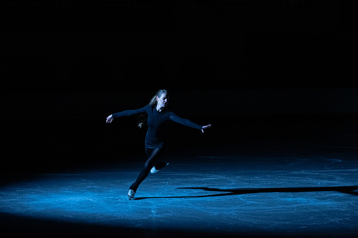 Professional figure skating artist slides and spins on the ice in the arena in the rays of blue light. Young woman practicing repertoire of an ice show. Artistic sport, grace, precision and perfection.