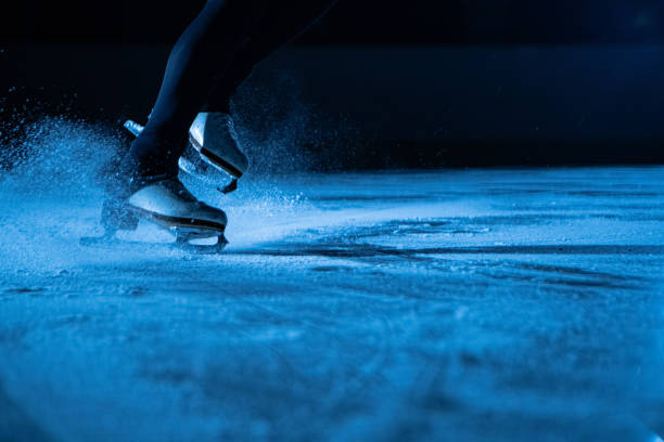 detaillierte aufnahme von frauenbeinen in weißen eiskunstlaufschlittschuhen auf kalter eisarena im dunkeln mit blauem licht. eine frau rutscht auf dem eis und spritzt glitzernde eispartikel in die kamera. aufschließen - eislaufen stock-fotos und bilder