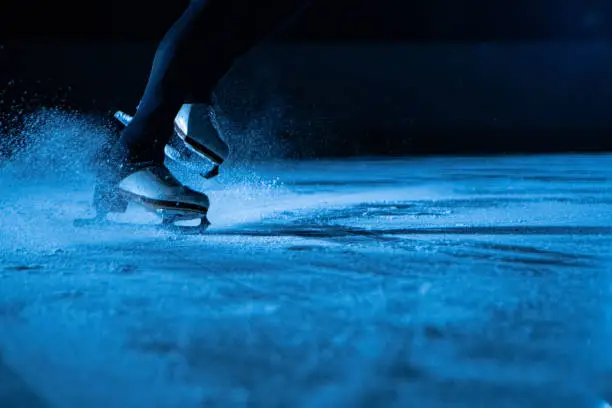 Detailed shot of women's legs in white figure skating skates on cold ice arena in the dark with blue light. A woman slides on the ice, splashing particles of sparkling ice into the camera. Close up