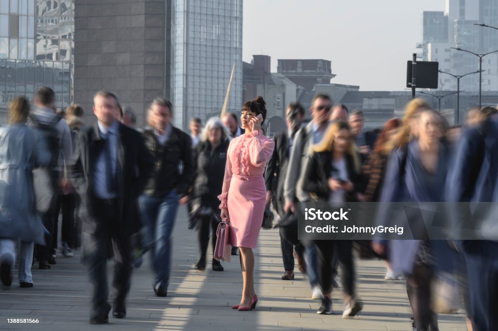 Executive dressed in pink using phone during morning commute Three-quarter front view of smiling businesswoman with briefcase stopped on London Bridge while walking with crowd from Southwark to financial district. Individuality Stock Photo