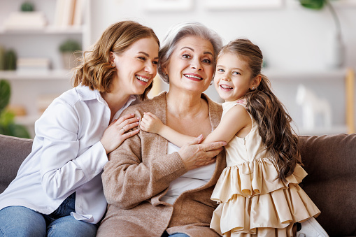 Happy multi generational family: elderly female hugging little girl and young woman smiling  while sitting on sofa together