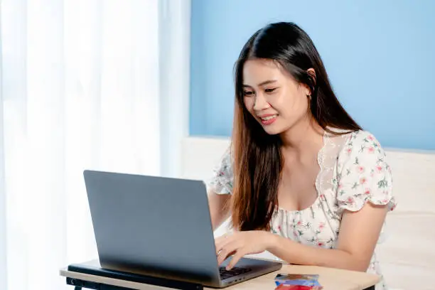 Beautiful Asian girl,sitting in front of her laptop screen, shopping and ordering online, with a smiling and happy face, on the table are several credit cards,online shopping.
