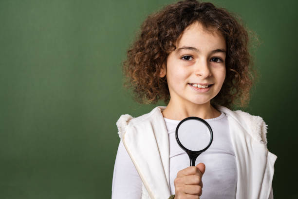 one small caucasian girl ten years old with curly hair front view portrait close up standing in front of green background looking to the camera holding magnifying lens education and learning concept - magnifying glass lens holding europe imagens e fotografias de stock