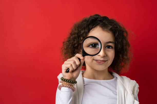 one small caucasian girl ten years old with curly hair front view portrait close up standing in front of red background looking to the camera holding magnifying lens education and learning concept - magnifying glass lens holding europe imagens e fotografias de stock