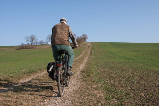 An elderly man with his bicycle rides on a field path on a beautiful spring day between the fields up a hill towards the blue sky.