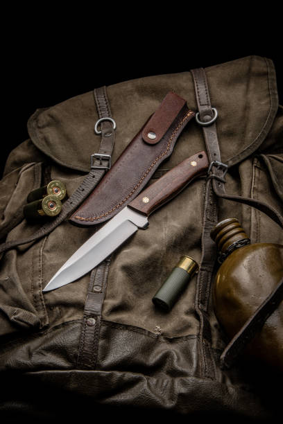 istock Hunting knife with a wooden handle on a vintage canvas backpack. 12-caliber shotgun shells and a water canteen are nearby. Hunter's set. Dark background. 1388627178