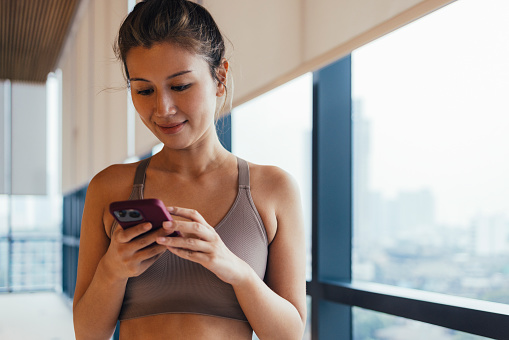 Asian female in her 30s texting on her smartphone after a workout in the gym.