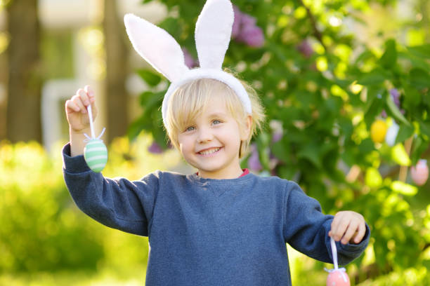 Pretty little boy wearing bunny ears hunting for eggs in spring park on Easter day.Traditional festival outdoors. stock photo