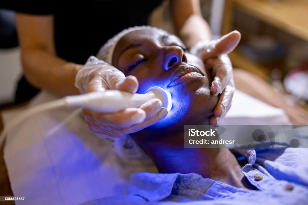 Woman at the spa getting a rejuvenation treatment on her face African American woman at the spa getting a rejuvenation treatment on her face using ultrasound and light therapy Facial Mask - Beauty Product Stock Photo