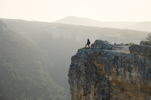 Tourist cliff mountain. Silhouette against the background of bright sunlight. A young man stands on the edge of a cliff. The concept of an active lifestyle, the courage of brave decisions. Travel