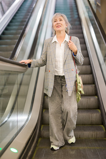 Positive senior lady walking down through escalator to metro station.