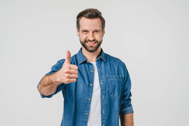 portrait en gros plan d’un jeune homme de race blanche positif en vêtements décontractés en jean montrant le pouce levé pour un bilan de bonne qualité, souriant avec un sourire aux dents isolé dans un fond blanc - toothy smile relaxation white healthcare and medicine photos et images de collection