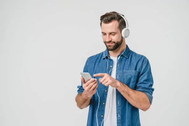 Photo of Caucasian young student man freelancer listening to the music in headphones, choosing sound track, song, playlist, podcast on phone isolated in white background
