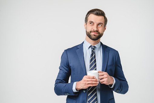 Thoughtful pensive caucasian young handsome successful businessman in formalwear holding cup of hot beverage tea coffee looking at the side thinking isolated in white background