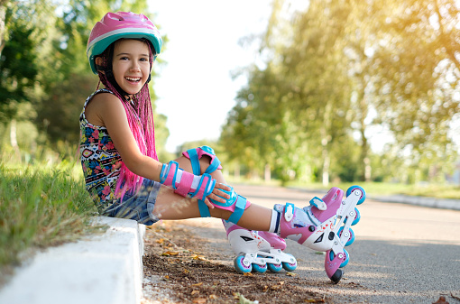 Laughing loudly, a funny girl with Afro-pigtails and wearing sports protective gloves and a helmet sat down to rest on the sidewalk after roller skating. A child's favorite hobby is roller skates.