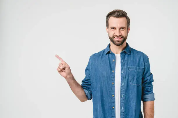 Happy caucasian young man in denim casual clothes pointing at copy space showing freespace isolated in white background. Ad place