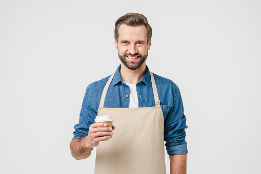Smiling caucasian young man barista coffee maker bartender holding hot beverage tea paper cup wearing apron isolated in white background