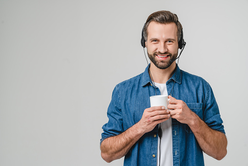 Successful caucasian male it support hotline worker in headphones drinking coffee hot beverage helping clients on calls isolated in white background