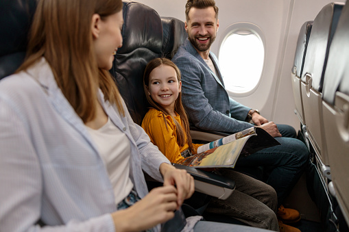 Joyful child holding magazine and smiling while sitting in passenger chair next to father and mother in aircraft