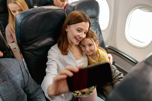Smiling woman hugging child and taking picture with smartphone while sitting in passenger chair during flight