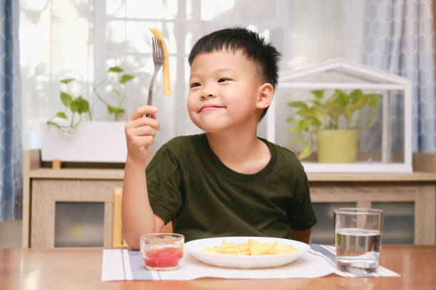 Happy little Asian kindergarten boy child using fork eating French fries potato chips with ketchup stock photo
