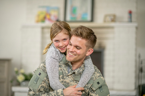 A father and young daughter are embracing in their living room. The dad is wearing a military uniform and has just returned home.