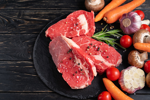 Three pieces of juicy raw beef with rosemary on a stone cutting board on a black wooden table background. Meat for barbecue or grill with vegetables pepper and salt seasoning