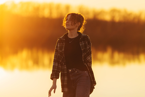 Redhead curly beautiful girl against sunset. Close up portrait. Spring break on river bank. Woman enjoying vacation. Visit local attractions. Girl in nature. Feel happiness. Local travel. Morning sun