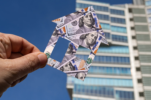 Symbolic house with the image of 100 American dollars in a man's hand against the background of a modern building
