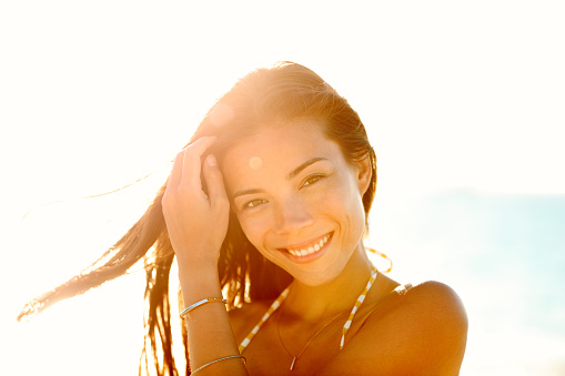 Beautiful happy Asian chinese woman in bikini touching her healthy soft hair in sunset flare at beach sunset on summer vacation travel. Multiracial model wearing gold jewelry bracelet and necklace.