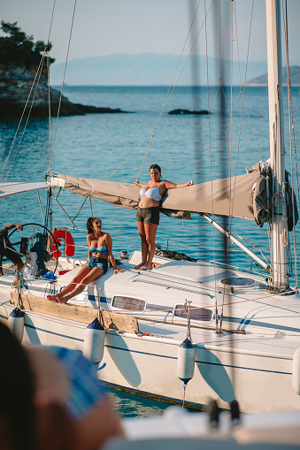 Two young women in bikini tops and shorts enjoying sunshine on sailing boat deck by the coastline, relaxing on summer vacation