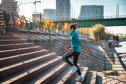 Young woman in sports clothes running up the stairs by the city river, residential buildings construction site in the background, healthy urban lifestyle