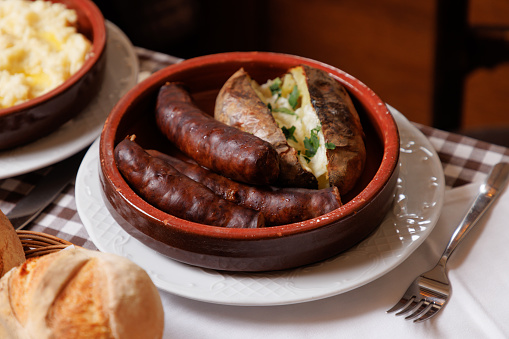 Smoked sausages served with whole baked potato cut in half in traditional crockery, bread buns in basket on ethnic restaurant table