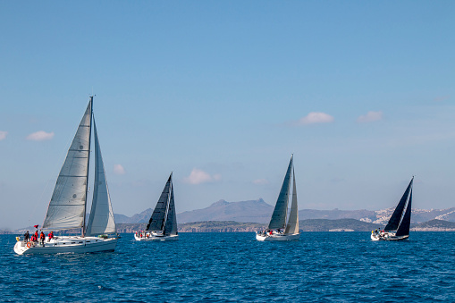 Sailboats sail in windy weather on blue waters of the Aegean Sea at the shores of famous holiday destination Bodrum, Turkey. Sailing crew racing at regatta. Yacht boat with mast. Water splashes, islands, cloudy sky and seascape background with copy space.