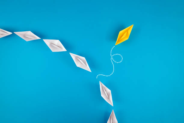 istock Group of White Paper Boats with One Yellow Boat Going Different Direction 1388725317