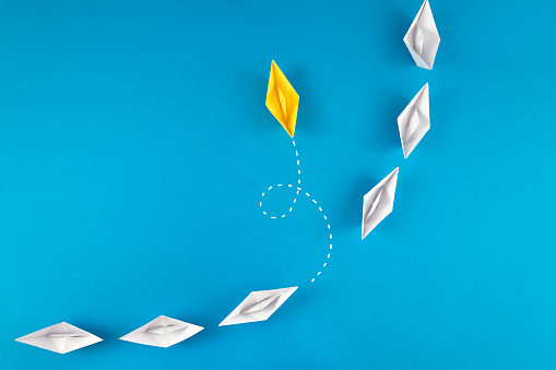 Group of white paper boats and one yellow paper boat going different direction on blue background