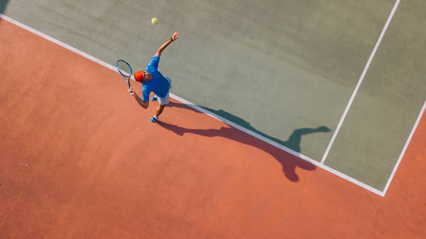 drohnen-blickwinkel asiatischer tennisspieler serviert den ball mit schatten direkt darüber - sportmatch stock-fotos und bilder