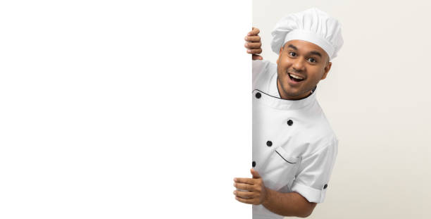 istock Happy Young handsome asian man chef in uniform looks out from behind an empty whiteboard. Cooking indian man hiding behind big blank billboard for advertising text menu in kitchen and restaurant. 1388729037