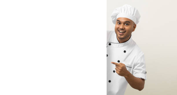 istock Happy Young handsome asian man chef in uniform looks out from behind an empty whiteboard. Cooking indian man hiding behind big blank billboard for advertising text menu in kitchen and restaurant. 1388729611