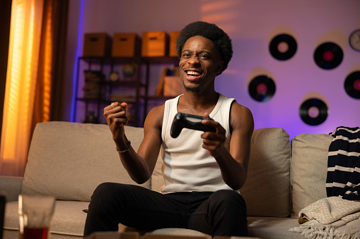 A male athlete wins a round in a console game. The man is sitting on the couch in front of the TV with the pad in hand, clenching fist with joy, smiling