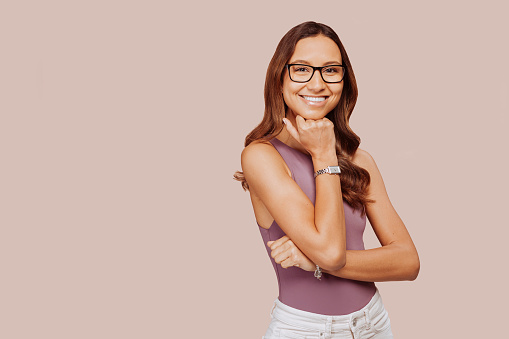 Cheerful young stylish businesswoman wears eyeglasses, hold hand on chin look at camera isolated on beige background. Confident multiracial female student professional portrait. Copy space.