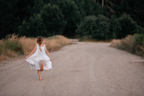 A little barefoot girl in a long white dress runs along the sandy road of a wide road.Summer walk in the countryside. Carefree childhood.