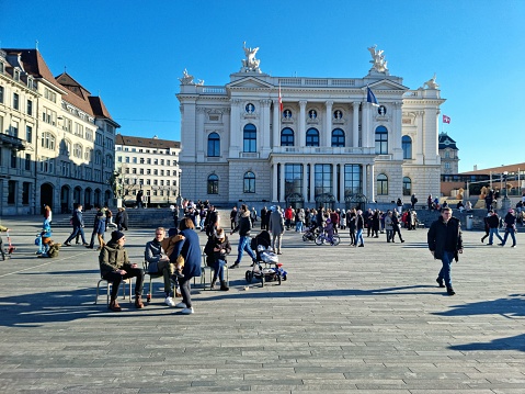 The Opera Theater building in Zurich City during a beautiful day in spring season. The image shows the Sechseläuteplatz in front of the Opera with many pedestrians going out for a walk.