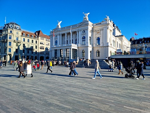 The Opera Theater building in Zurich City during a beautiful day in spring season. The image shows the Sechseläuteplatz in front of the Opera with many pedestrians going out for a walk.