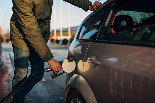 A man in blue jeans and a winter jacket holds a pump that fills gasoline in the tank