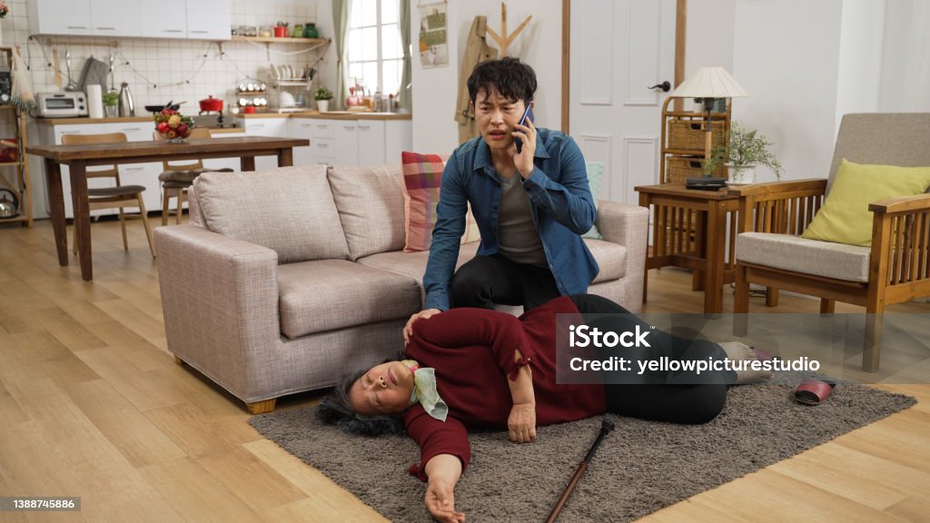worried Asian adult man son talking to emergency service call on the phone beside his unconscious mother lying on the ground after a fall at home. Falling Stock Photo