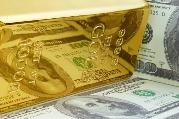 Yellow gold ingot bar with reflection of dollar bill prop paper money.